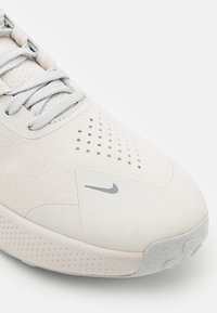 Scarpa sportiva grigia chiara con una tomaia testurizzata, perforazioni per la traspirabilità e una suola in gomma con un piccolo logo Nike.