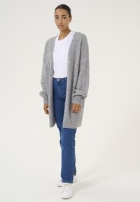 Grauer, gestrickter Cardigan mit langen Ärmeln, offenem Vorderteil und geripptem Muster, kombiniert mit einem weißen T-Shirt und blauen, geraden Jeans. Weiße Sneaker.