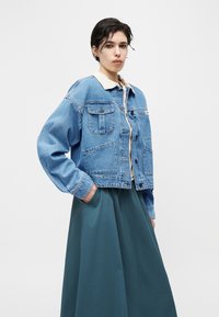 Νεαρό άτομο που φορά ανοιχτό γαλάζιο τζιν μπουφάν πάνω από λευκό πουκάμισο και σκούρα τυρκουάζ φούστα, στέκεται με τα χέρια στις τσέπες μπροστά σε απλό φόντο.