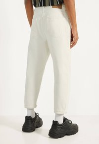 Bílé džínové kalhoty s ohrnutými lemy, dvěma zadními kapsami a koženou nášivkou na pasu, doplněné černými robustními teniskami.