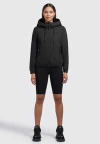 Veste à capuche noire avec fermeture éclair, poignets élastiques et poches latérales. Portée sur un short de cyclisme noir et des baskets noires. Texture de tissu lisse.