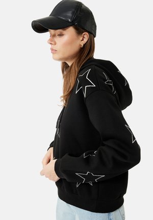 Kvinde iført sort hættetrøje med hvide stjernedesigns og sort læderkasket, stående i profil med hænderne ved taljen.