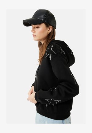 Vrouw die een zwarte hoodie met witte sterontwerpen en een zwarte leren pet draagt, in profiel staand met handen bij de taille.