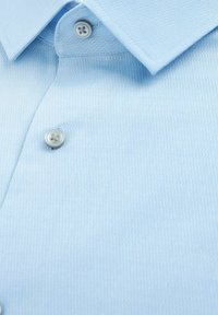 Hellblaues kurzärmliges Hemd aus strukturiertem Stoff, mit einem spitzen Kragen und zwei grauen Knöpfen. Nahaufnahme des Materials.