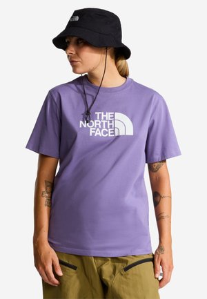 Persoon draagt een paarse The North Face t-shirt, olijfgroene broek met zwarte ritsen en een zwarte bucket hat met kinbandjes.