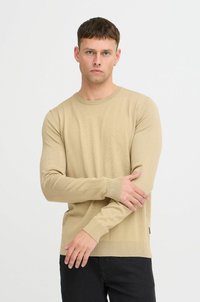 Suéter de manga larga beige claro con cuello redondo. Textura suave, diseño ajustado, puños y dobladillo acanalados. Combinado con jeans oscuros.