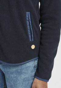 Veste en polaire de marine avec une surface texturée, dotée d'une poche d'accent bleue et d'un détail de logo circulaire à l'ourlet.