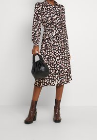 Vestido de estampado de leopardo con mangas largas, cierre de botones en la parte delantera y cinta en la cintura. Presenta un bolso negro arrugado y botas de tacón marrones.