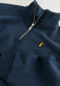 Sweatshirt zippé marine avec un col haut, des poignets côtelés et un logo de homard brodé jaune sur la poitrine. Tissu doux et texturé.