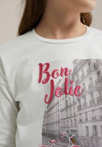 Balta medvilninė marškinėlių su spausdintu miesto peizažu ir rožinėmis siuvinėtomis raidėmis "Bon Jolie". Detalės apima dviratį su krepšiu ir gėlėmis.