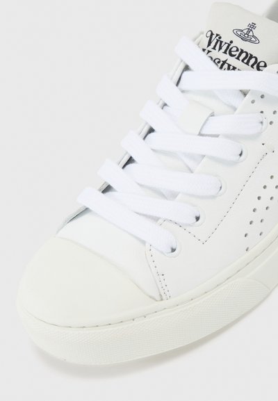 Valkoinen nahkainen lenkkari, jossa on rei'itetty yksityiskohta, litteät valkoiset nauhat ja rakennekumipohja. Ominaisuuksiin kuuluu pyöreä kärki ja logo kielen kohdalla.