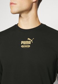 Felpa nera con colletto a costine. Presenta un logo dorato "PUMA KING" e una grafica di puma sul petto. Il materiale sembra morbido.