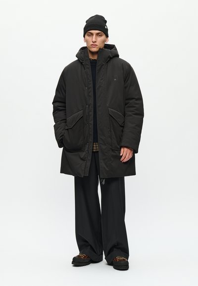 Man wearing black padded hooded coat, black beanie, wide black trousers, and brown shoes standing against white background.
