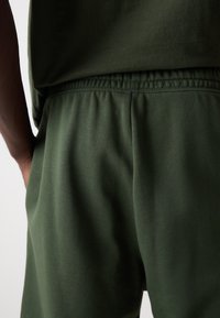 Mørkegrønne bomuldsshorts med elastisk talje, der har en glat tekstur og en løs pasform, delvist skjult af en matchende top.