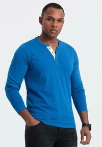 Mies, joka käyttää sinistä pitkähihalaista henley-paitaa, jossa on valkoinen napitus, mustia farkkuja ja mustaa rannekelloa, seisoo yksi käsi taskussa.