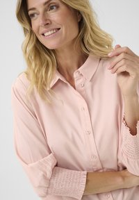 Blusa rosa chiaro con colletto classico, chiusura frontale con bottoni e polsini arricciati. Tessuto liscio con una finitura leggermente lucida.