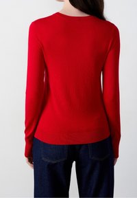 Red long-sleeve top made from soft fabric, featuring a rounded neckline and ribbed hem. Paired with dark blue jeans, showing back view.