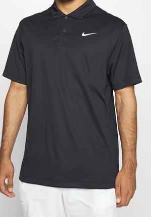 Homme portant un polo noir Nike à manches courtes avec deux boutons, associé à un pantalon blanc, vu des épaules jusqu'à la mi-cuisse.