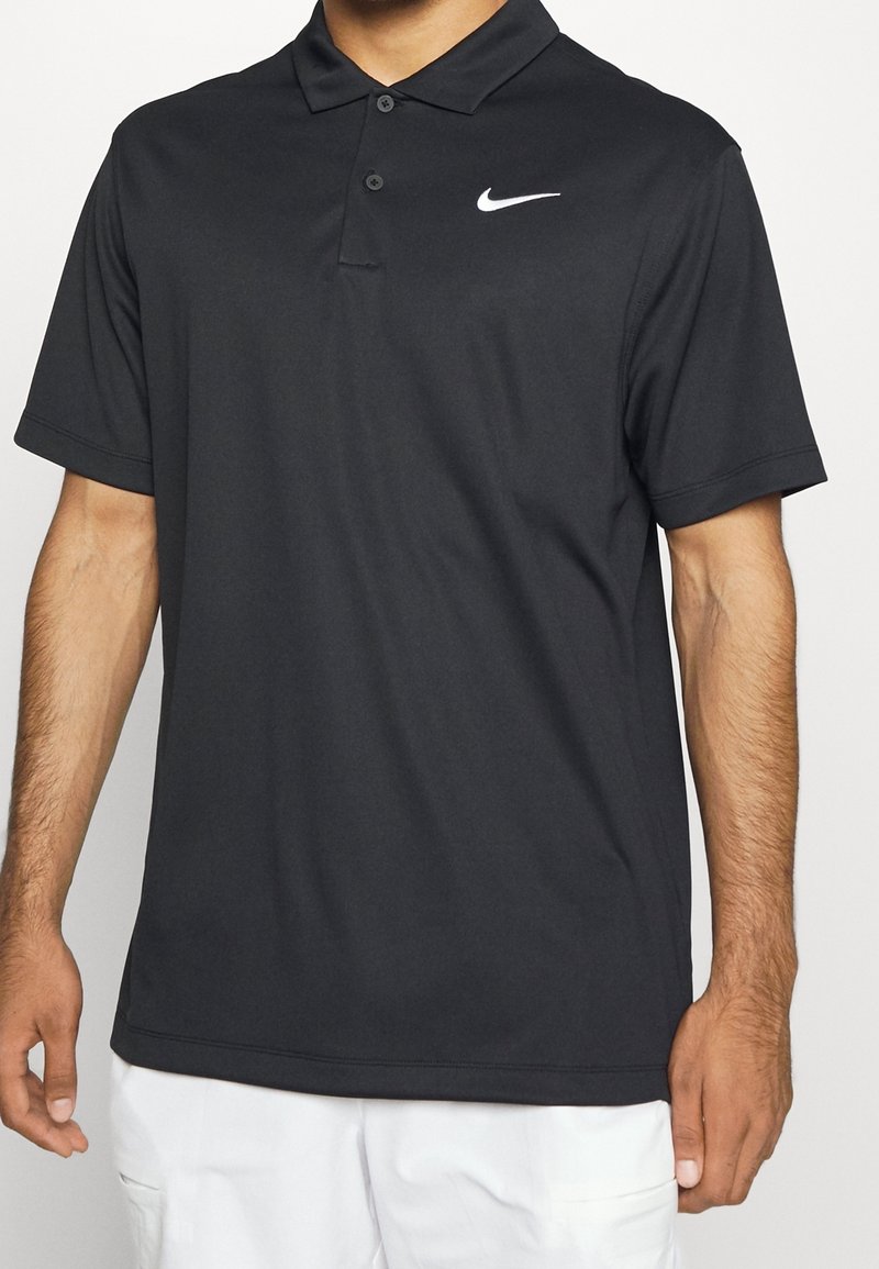 Homme portant un polo noir Nike à manches courtes avec deux boutons, associé à un pantalon blanc, vu des épaules jusqu'à la mi-cuisse.