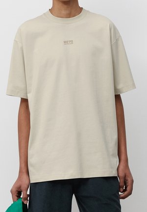 Osoba nosząca luźny beżowy T-shirt z napisem "MO'PD" umieszczonym na środku klatki piersiowej, trzymająca zielony przedmiot w lewej ręce.