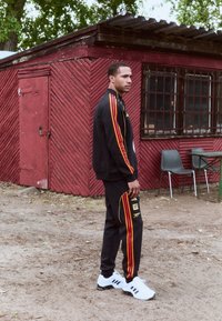 Black athletic tracksuit with red and orange stripes, featuring a logo. White athletic shoes with black accents, standing on sandy ground.