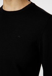 Svart stickad tröja med rund halsringning och en liten logotypdetalj på bröstet. Texturen ser mjuk och lätt ut.