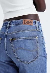Jean en denim bleu avec des coutures jaunes, featuring an embossed logo patch in brown leather at the back of the waistband and two poches arrière.