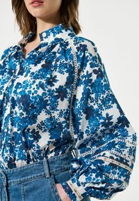 Langdurige katoenen blouse met lange mouwen in het wit, bedrukt met een blauw bloemenpatroon. De blouse heeft gathered mouwen en contrasterende decoratieve afwerking.