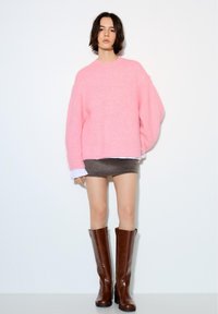 Femeie purtând un pulover roz pufoș, fustă mini gri și cizme maro din piele până la genunchi, stând în fața unui fundal alb.