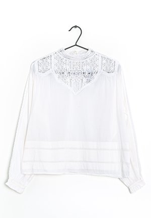 Hvid bluse med lange ærmer, blonderyg, høj halsudskæring og delikat kant på ærmerne, hængende på en sort bøjle mod hvid baggrund.