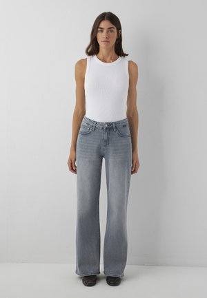 Woman with dark hair wearing a sleeveless white top, light gray wide-leg jeans, and black shoes standing against a plain light gray wall.