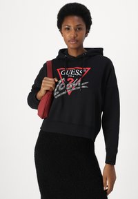 Felpa nera corta con un logo triangolare rosso e testo bianco. Abbinata a una gonna nera texture e con in mano una piccola borsa rossa.