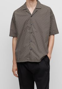 Camisa de manga corta de tejido marrón con un patrón geométrico en blanco y azul claro. Presenta un cuello y un cierre de botones al frente.