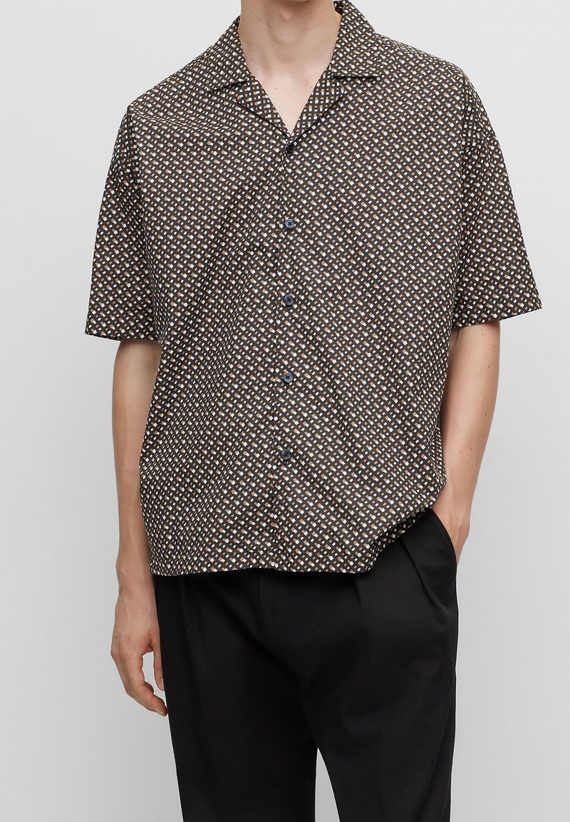 Camisa de manga corta de tejido marrón con un patrón geométrico en blanco y azul claro. Presenta un cuello y un cierre de botones al frente.