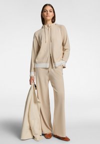 Completo sportivo beige con giacca a zip con cappuccio, polsini a righe e pantaloni a gamba larga. Tessuto liscio con una vestibilità ampia. Scarpe marroni.