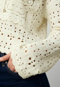 Pull en crochet couleur crème avec des motifs de trous complexes, des bords festonnés et des manches larges, associé à un jean en denim foncé.