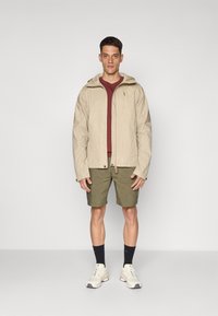 Beige hettejakke med glidelås, lagd over en bordeaux skjorte, kombinert med olivengrønne shorts, hvite joggesko og mørke sokker.