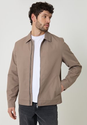 Mand iført en beige lynlåstrøje over en hvid t-shirt og mørke jeans, kigger mod højre med den ene hånd i lommen.