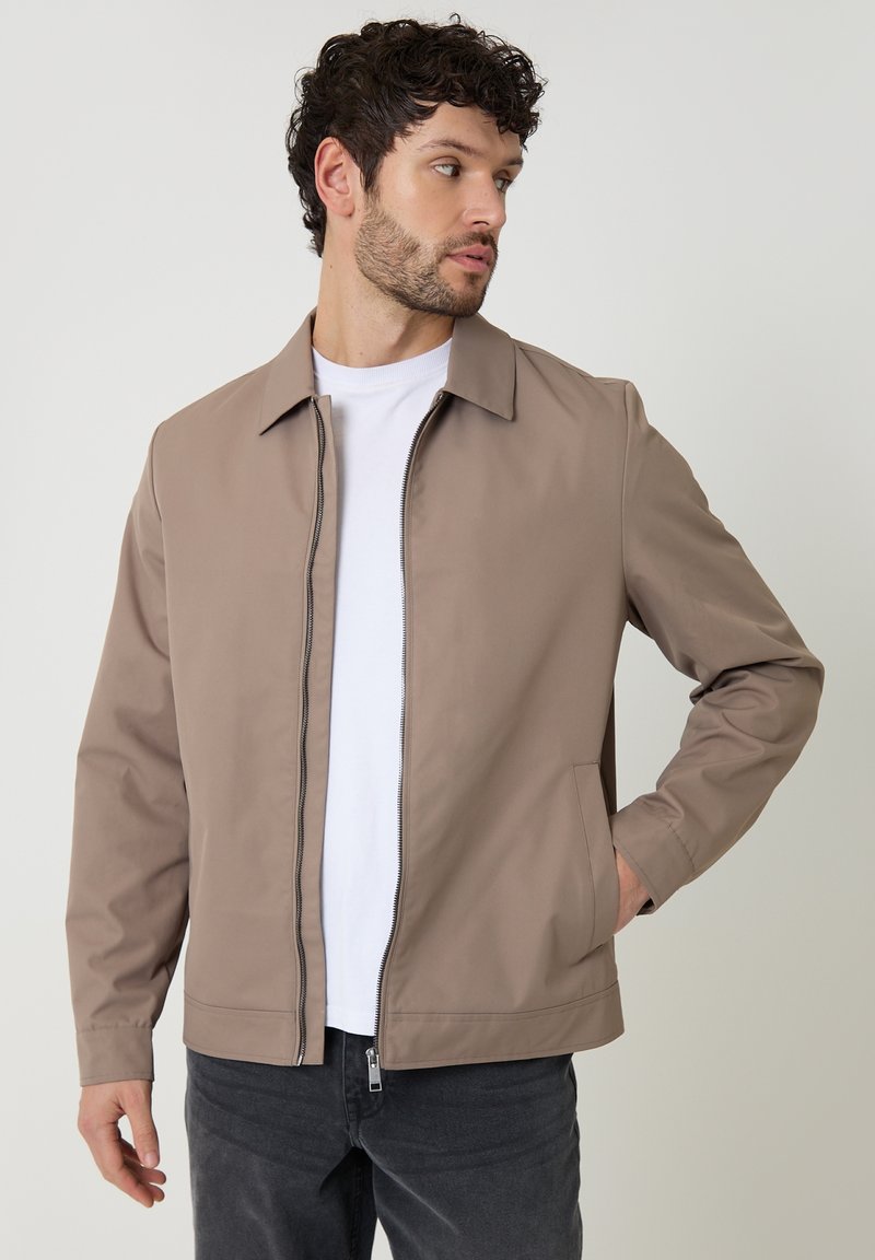 Man draagt een beige jack met ritssluiting over een wit T-shirt en donkere spijkerbroek, kijkt naar rechts met één hand in zijn zak.