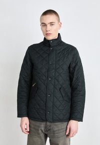 Černá prošívaná bunda s vysokým límcem, kapsami na zip na přední straně a patentkami. Měkká textura, minimalistický design.