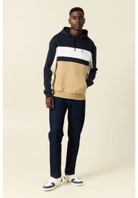 Felpa con cappuccio in navy, bianco e beige con tasca frontale. Abbinata a jeans denim scuri e sneakers bianche con dettagli blu.