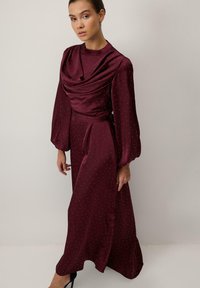 Egy nő hosszú bordó színű ruhát visel, amely ezüst pöttyökkel díszített, hosszú puffos ujja van, és drapált dekoltázsú. Egy világos sima fal előtt áll.