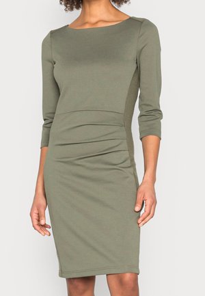 Olśniewająca oliwkowozielona sukienka o dopasowanym kroju do kolan, z rękawami do łokcia i subtelnymi marszczeniami z boku, na stojącej osobie.