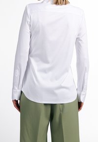 Persona che indossa una camicia bianca a maniche lunghe aderente con polsini abbottonati e pantaloni larghi verde oliva, vista da dietro.