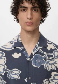 Camicia a maniche corte di colore blu navy con grandi motivi floreali bianchi. Presenta un colletto a camicia e chiusura con bottoni. Tessuto morbido e strutturato.