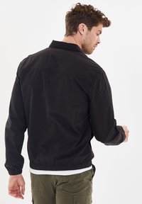 Zwarte jas van katoen, met lange mouwen, een kraag en een elastische taille. De achterkant toont een gladde textuur en geen zichtbare versieringen.
