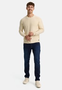 Chemise beige clair à manches longues avec un col rond, assortie à un jean en denim foncé et des baskets beiges, se tenant devant un fond blanc.