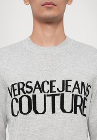 Pelēks džemperis ar melnu adītu uzrakstu "VERSACE JEANS COUTURE." Apaļš kakls un ribotās aproces, mīksta tekstūra. Klasiska silueta dizains.