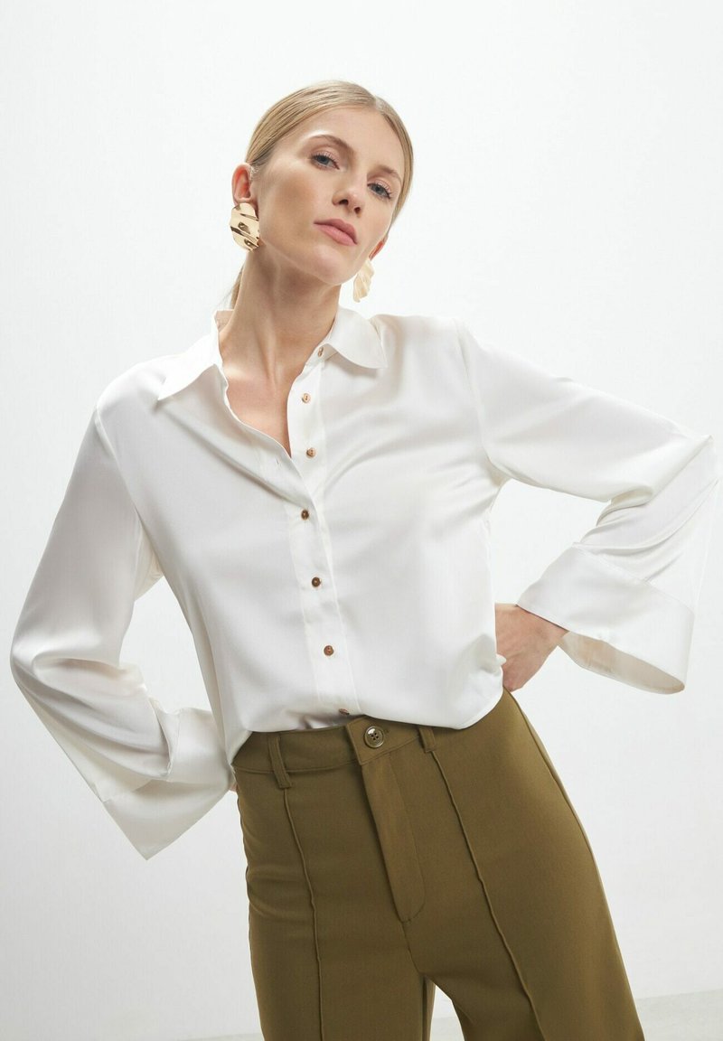 Blouse en satin blanc avec des manches longues en forme de cloche et un design à boutons, associée à un pantalon taille haute vert olive. Des accessoires en or complètent la tenue.