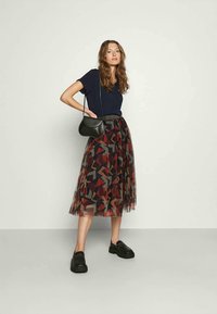 Mulher de pé vestindo uma camisa azul-marinho, uma saia midi estampada, mocassins pretos e uma pequena bolsa transversal preta, contra um fundo claro e liso.
